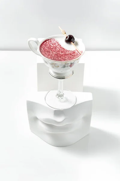 Rock Island cocktail with raspberry powder and grapes on a skewer. A cocktail in a glass in the shape of a cup on a long stem stands on a plaster figure among plaster figures on a white background.