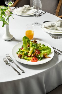 Hafif seramik tabakta sebze salatası, kızarmış enginar, ramen yaprağı, domates ve parmesan. Tabak beyaz bir masa örtüsünün üzerinde duruyor, yanında bir bardak ve alkolsüz şarap ve çatal bıçak takımı var..