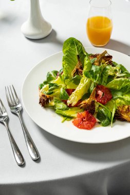 Hafif seramik tabakta sebze salatası, kızarmış enginar, ramen yaprağı, domates ve parmesan. Tabak beyaz bir masa örtüsünün üzerinde duruyor, yanında bir bardak ve alkolsüz şarap ve çatal bıçak takımı var..