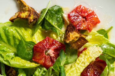 Hafif seramik tabakta sebze salatası, kızarmış enginar, ramen yaprağı, domates ve parmesan. Tabak beyaz bir masa örtüsünün üzerinde duruyor, yanında bir bardak ve alkolsüz şarap ve çatal bıçak takımı var..