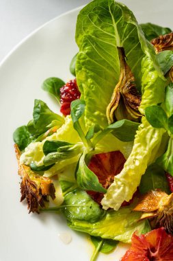 Hafif seramik tabakta sebze salatası, kızarmış enginar, ramen yaprağı, domates ve parmesan. Tabak beyaz bir masa örtüsünün üzerinde duruyor, yanında bir bardak ve alkolsüz şarap ve çatal bıçak takımı var..