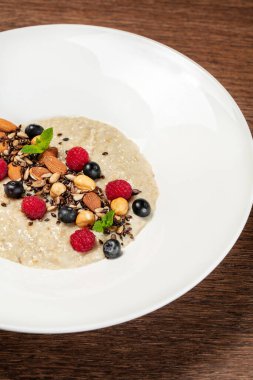 Çeşit çeşit fındıklı yulaf ezmesi, ahududu ve nane yapraklı yaban mersini seramik tabakta. Tabak, portakal suyu olan bir bardağın yanında, ahşap bir masanın üzerinde duruyor..