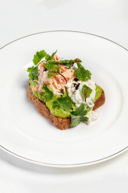 Haşlanmış yumurta, avokado püresi, marul ve yengeç etli maydanoz. Hafif seramik tabakta yemek, masa örtüsü olan bir masada. Yakınlarda bir vazo ve çatal bıçak takımı var..