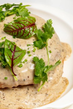 Mantar soslu jambon ve peynirli Brioche çöreği. Üstü marul, maydanoz ve doğranmış yeşil soğanla kaplı. Hafif bir masa örtüsü üzerinde hafif bir tabakta yemek. Yakınlarda bir fincan çay ve çaydanlık var..