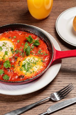 İki yumurtalı Shakshuka, üzerinde siyah bir tavada doğranmış otlar ve sapı olan. Yakınlarda bir çay tabağı ve portakal suyu var. Tabaklar kahverengi ahşap bir masada..