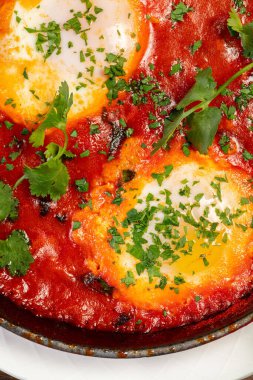 İki yumurtalı Shakshuka, üzerinde siyah bir tavada doğranmış otlar ve sapı olan. Yakınlarda bir çay tabağı ve portakal suyu var. Tabaklar kahverengi ahşap bir masada..