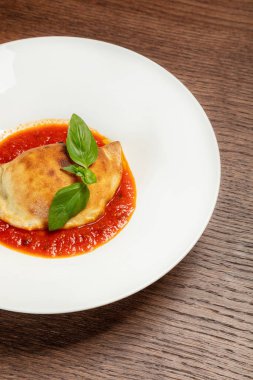 Domates sosu ve fesleğen yapraklarıyla kaplı mozzarella peynirli mini fırın fırını. Yiyecek, kahverengi bir masanın üstündeki açık seramik tabakta yatıyor..