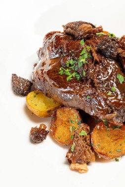 Şarap soslu dana yanakları, tepesinde doğranmış otlar ve hafif seramik tabakta karyolada kızarmış patatesler. Tabak kahverengi, ahşap bir tezgahta duruyor..