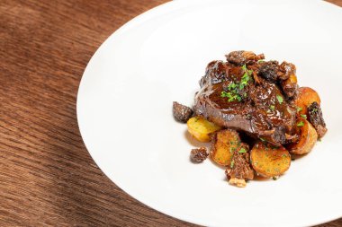 Şarap soslu dana yanakları, tepesinde doğranmış otlar ve hafif seramik tabakta karyolada kızarmış patatesler. Tabak kahverengi, ahşap bir tezgahta duruyor..