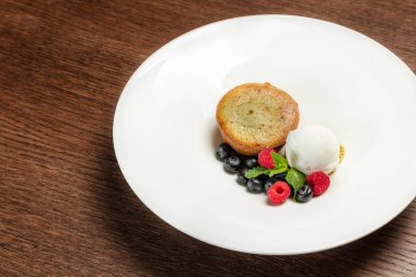 İçinde şam fıstığı kremalı beyaz çikolata, vanilyalı dondurma, şam fıstıklı cips, yabanmersini ve ahududu, yakınlarda nane yaprakları. Tahta bir masanın tepesinde hafif bir tabakta tatlı.