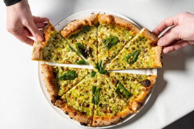 Ekşi hamurlu pizza, dört peynirli jalapeno ve şam fıstığı ve hafif seramik tabakta parmesan peyniri. Tabak hafif bir masa örtüsünün üzerinde duruyor ve iki el farklı taraflardan bir parça alıyor..