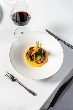 Kırmızı şarapta kızartılmış ciğer, haşlanmış sebzeler ve geniş kenarlı beyaz tabakta kabak püresi. Tabak, kırmızı şarap ve çatal bıçak takımının yanında beyaz bir masa örtüsünün üzerinde duruyor..