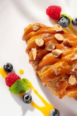 Üstünde karamel ve fındıklı Fransız kruvasanı, yanında da ahududu ve nane yapraklı yaban mersini. Hafif bir tabakta yemek, masa örtüsü ve yakınlarda bir fincan kahve..