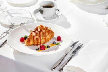 Üstünde karamel ve fındıklı Fransız kruvasanı, yanında da ahududu ve nane yapraklı yaban mersini. Hafif bir tabakta yemek, masa örtüsü ve yakınlarda bir fincan kahve..