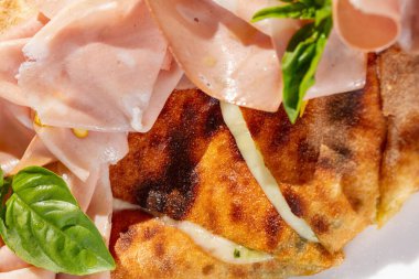 Peynirli Calzone pizza, üstüne de jambon ve fesleğen dilimleri. Tabak hafif bir tabakta yatıyor. Taoelka masa örtüsünün üzerinde duruyor, yanında beyaz şarap ve çatal bıçak takımı var..