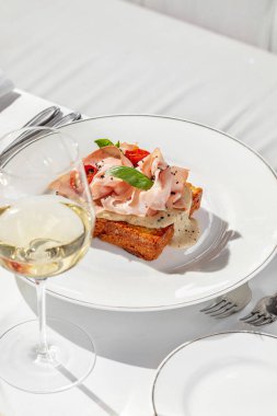 Jambonlu Brioche tostu ve fesleğen yaprağı ve bitkilerle kaplı ceviz sosu. Tost hafif bir seramik tabakta, yanında bir bardak beyaz şarap var..