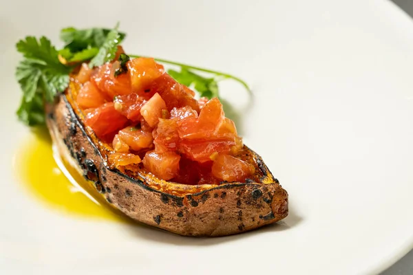 烤红薯 顶部有新鲜的切碎的西红柿 在薄薄的瓷盘里放上一小枝橄榄油香菜 盘子放在桌布上 旁边是油瓶和餐具 — 图库照片
