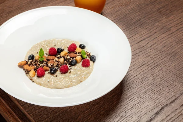 Stock image Oatmeal with nuts of various kinds, raspberries and blueberries with mint leaves in a light ceramic plate. The dish stands on a wooden table, next to a glass with orange juice.