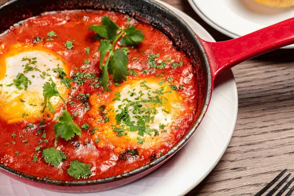 stock image Two egg shakshuka topped with chopped herbs in a black frying pan with a handle. Nearby is a saucer with a bun and orange juice. Dishes are on a brown wooden table.