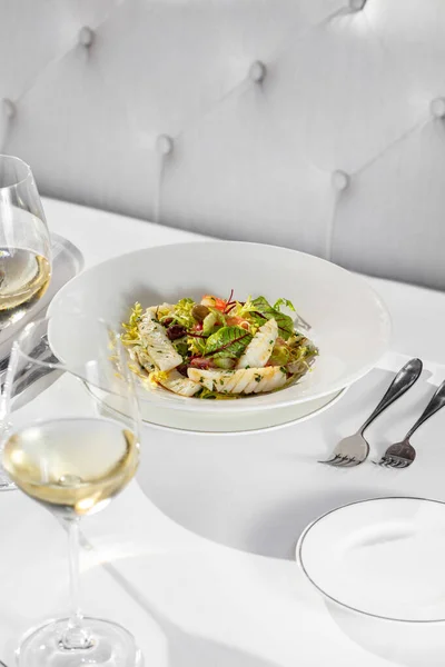 白ワインソースにパプリカとトラウトフィレを混ぜたサラダ 皿は薄い陶板の中にある 皿はテーブルクロスの上にあり その隣には白ワインとカトラリーのガラスがあります — ストック写真