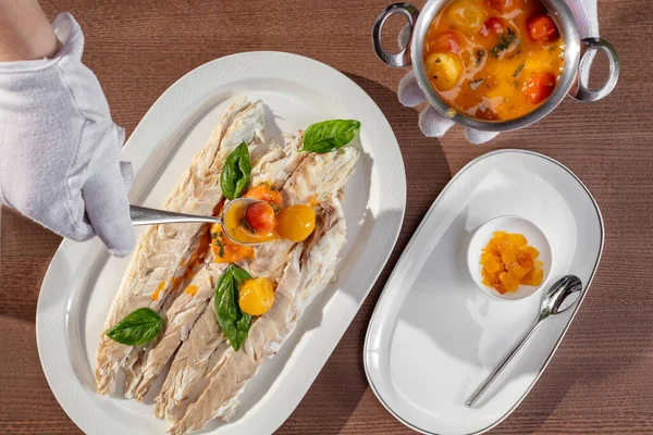 stock image A trout fillet lies on a flat ceramic plate on a brown table. Nearby is a dish with a metal bowl with tomatoes in wine sauce. The waiter spoons the tomatoes in the sauce onto the trout fillet.