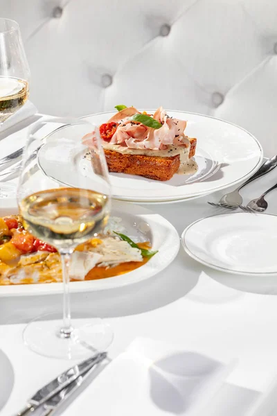 ハムとトマトのくるみソースとブリオッシュトースト バジルの葉とハーブをトッピング トーストは薄いセラミックプレートの上にあり 白ワインのグラスとマスの入った料理が近くにあります — ストック写真