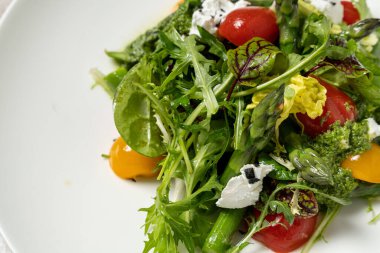 Kuşkonmaz soslu bahar salatası, kırmızı ve sarı kiraz domatesleri, garganzola peyniri, marul karışımı ve pesto soslu mikroçerez. Salata, kumaş arka planında hafif bir seramik tabakta yatıyor..