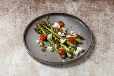 Izgarada keçi peyniri ve balzamik soslu domatesli taze Ukrayna kuşkonmazı. Yiyecek, kumaş arka planında, siyah bir tabakta yatıyor..