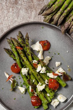 Izgarada keçi peyniri ve balzamik soslu domatesli taze Ukrayna kuşkonmazı. Yiyecek, kumaş arka planında, siyah bir tabakta yatıyor..