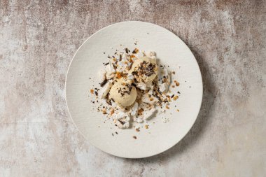 İki kepçe kahve aromalı dondurma içinde krema, çikolata cipsi ve fındıklı karamel. Tatlı beyaz bir seramik tabakta açık kahve renkli bir arka planda yatıyor..