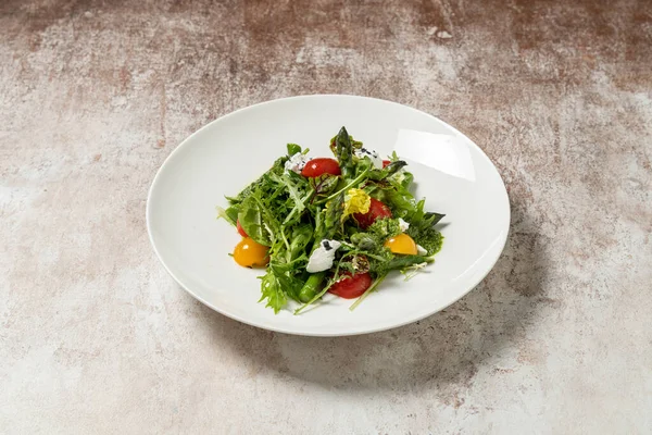 Kuşkonmaz soslu bahar salatası, kırmızı ve sarı kiraz domatesleri, garganzola peyniri, marul karışımı ve pesto soslu mikroçerez. Salata, kumaş arka planında hafif bir seramik tabakta yatıyor..