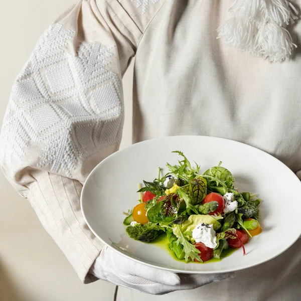 Spring salad with asparagus, red and yellow cherry tomatoes, garganzola cheese, lettuce mix and microgreens in pesto sauce. Salad in a light ceramic plate, in the hands of a girl in a Ukrainian vishyvanka