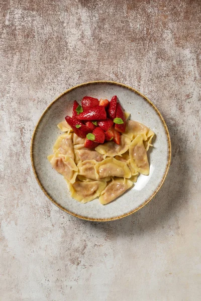 stock image Vareniki with cottage cheese and strawberries and slices of fresh strawberries on top. The dish lies in a light ceramic plate. The plate stands on a light, fabric background.