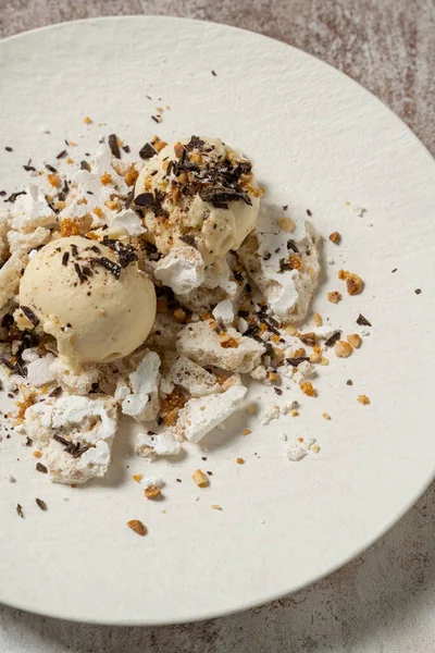 Two scoops of coffee flavored ice cream in a bowl with meringue, chocolate chips and caramel with hazelnuts. The dessert lies on a white ceramic plate on a light coffee-colored background.