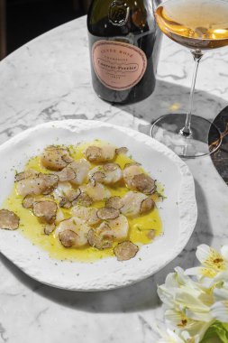 Mantarlı mantar dilimleri ve zeytinyağı ve bitkilerle karides carpaccio. Yakınlarda bir bardak köpüklü şarap ve bir şişe şarap var. Açık seramik tabakta mermer tezgahta yemek..