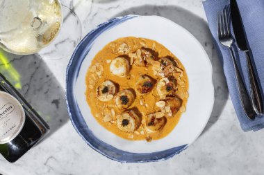 Grilled sea scallop with black caviar in cheese sauce with almond chips. The food lies in a light ceramic plate on a marble countertop. Nearby is a glass of white wine and a bottle of wine.