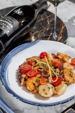 Deniz mahsullü makarna. Deniz tarağı, ahtapot, karides ve kremalı bitki soslu kiraz domatesli ev yapımı makarna. Makarna yuvarlak, hafif seramik tabakta yatıyor. Tabak hafif bir mermer masanın üzerinde duruyor. Yanında bir bardak Prosecco, keten peçetede çatal bıçak takımı.