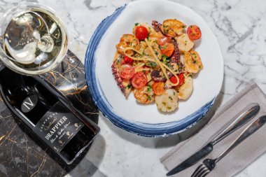 Deniz mahsullü makarna. Deniz tarağı, ahtapot, karides ve kremalı bitki soslu kiraz domatesli ev yapımı makarna. Makarna yuvarlak, hafif seramik tabakta yatıyor. Tabak hafif bir mermer masanın üzerinde duruyor. Yanında bir bardak Prosecco, keten peçetede çatal bıçak takımı.