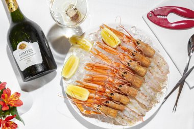 Italian dish. Shrimp Langoustine or Scampia, with lemon juice and olive oil with Italian spices. The shrimps lie on a pile of crushed crush ice, next to them lie slices of lemon. The food lies on a light, round, ceramic plate. The plate is on a table
