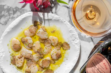 Mantarlı mantar dilimleri ve zeytinyağı ve bitkilerle karides carpaccio. Yakınlarda bir bardak köpüklü şarap ve bir şişe şarap var. Açık seramik tabakta mermer tezgahta yemek..