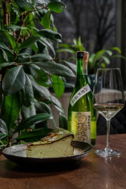 Fıstıklı kibrit peynirli kek ve seramik tabakta yeşil çay tozuyla kaplı hindistan cevizi cipsi. Masanın yanında bir bardak, bir şişe Japon şarabı ve bitkiler var..