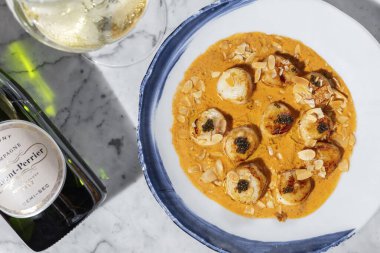 Grilled sea scallop with black caviar in cheese sauce with almond chips. The food lies in a light ceramic plate on a marble countertop. Nearby is a glass of white wine and a bottle of wine.
