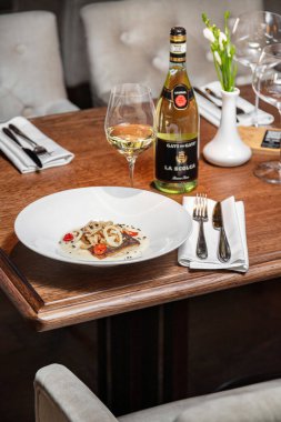 Hafif bir masa örtüsü üzerinde seramik tabakta patates püresi ve soğan cipsli domatesli tuzlu biftek. Yakınlarda bir bardak, bir şişe beyaz şarap ve çatal bıçak var..