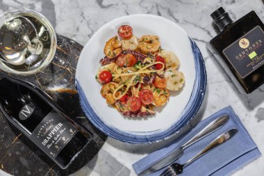 Deniz mahsullü makarna. Deniz tarağı, ahtapot, karides ve kremalı bitki soslu kiraz domatesli ev yapımı makarna. Makarna yuvarlak, hafif seramik tabakta yatıyor. Tabak hafif bir mermer masanın üzerinde duruyor. Yanında bir bardak Prosecco, keten peçetede çatal bıçak takımı.