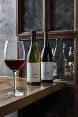 İki şişe kırmızı ve beyaz bulut şarabı ahşap, açık renkli bir masanın kenarında desenli duruyor. Yakınlarda kırmızı şarapla dolu uzun bir sapın üzerinde bir şarap kadehi duruyor. Masanın arkasında yansıyan şişelerin olduğu aynalı bir duvar var..