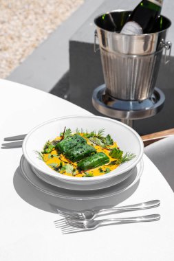 Krem peynirli somonla doldurulmuş ıspanaklı krep. Yemek seramik tabakta yatıyor. Tabaklar masanın üzerindeki hafif bir masa örtüsünün üzerinde, yanında da bir kova köpüklü şarap var..