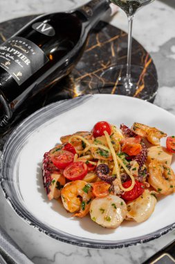 Deniz mahsullü makarna. Deniz tarağı, ahtapot, karides ve kremalı bitki soslu kiraz domatesli ev yapımı makarna. Makarna yuvarlak, hafif seramik tabakta yatıyor. Tabak hafif bir mermer masanın üzerinde duruyor. Yanında bir bardak Prosecco, keten peçetede çatal bıçak takımı.