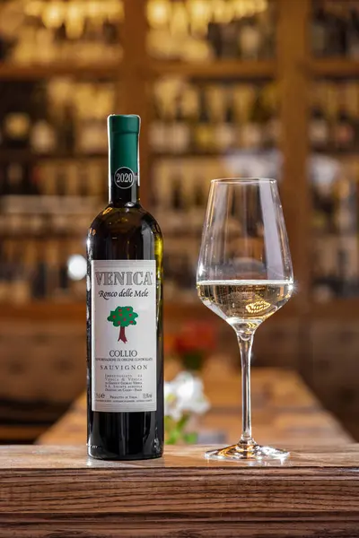 ソーヴィニヨン ロンコ ヴェニカ2021ワインのボトルは木製のスタンドに立っており その隣には高い茎に白ワインが入った透明なガラスが立っている ボトルとガラスの後ろには ワインのボトルが入ったカップボードがあります — ストック写真