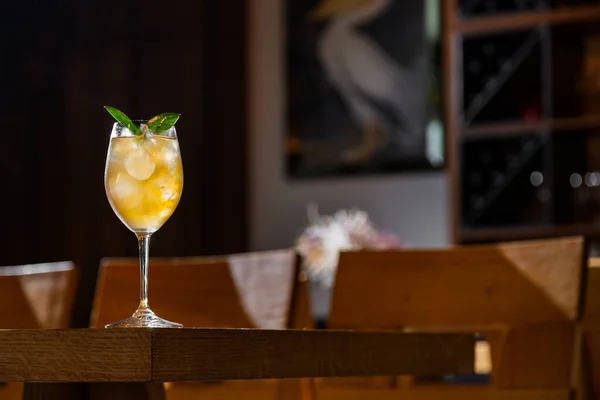 A cocktail with ice, syrup and basil in a glass that stands on the edge of a wooden table surrounded by a restaurant