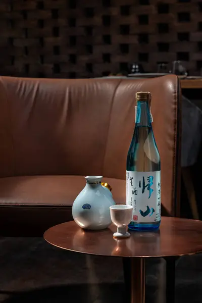 ショットグラスとデカンターを備えた日本酒のボトルは 茶色の革の椅子の隣の銅のテーブルに座っています — ストック写真